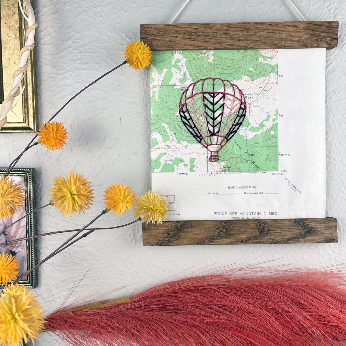 a vintage green and white USGS map of New Mexico hand embroidered with a hot air balloon in shades of mauve, framed on top and bottom with a rustic wood magnetic frame hanging on a grey wall with framed art and botanical decorations around it