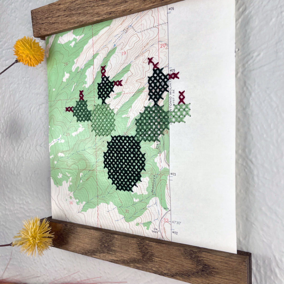 close up angled view of a vintage USGS map of New Mexico cross stitched with a prickly pear cactus, framed on top and bottom with a rustic wood magnetic frame hanging on a grey wall with botanical decorations around it
