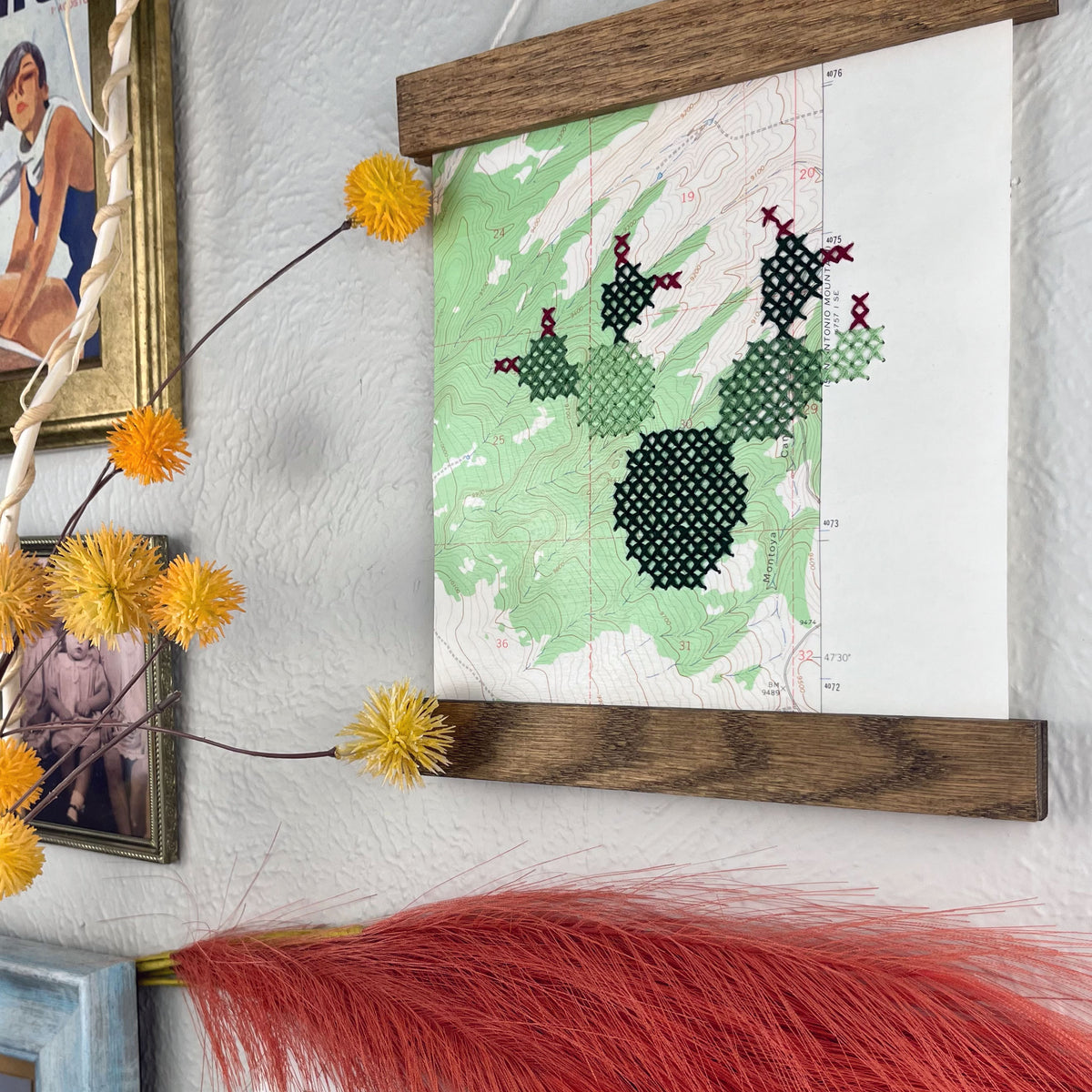 close up angled view of a vintage USGS map of New Mexico cross stitched with a prickly pear cactus, framed on top and bottom with a rustic wood magnetic frame hanging on a grey wall with botanical decorations and framed art around it