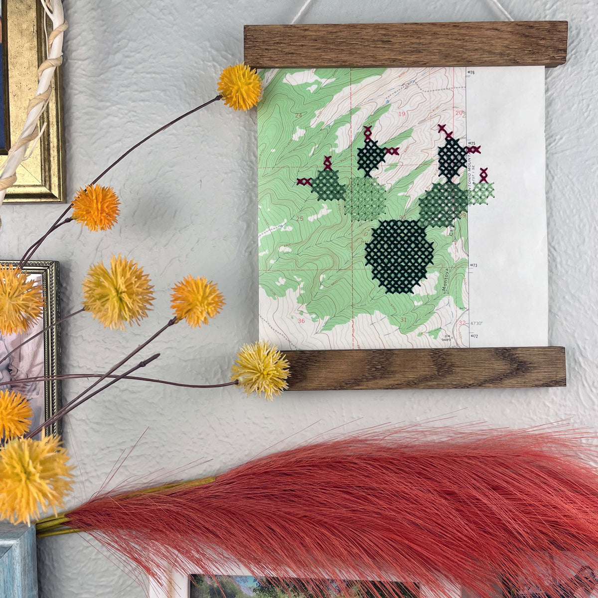 a vintage USGS map of New Mexico cross stitched with a prickly pear cactus, framed on top and bottom with a rustic wood magnetic frame hanging on a grey wall with botanical decorations and framed art around it