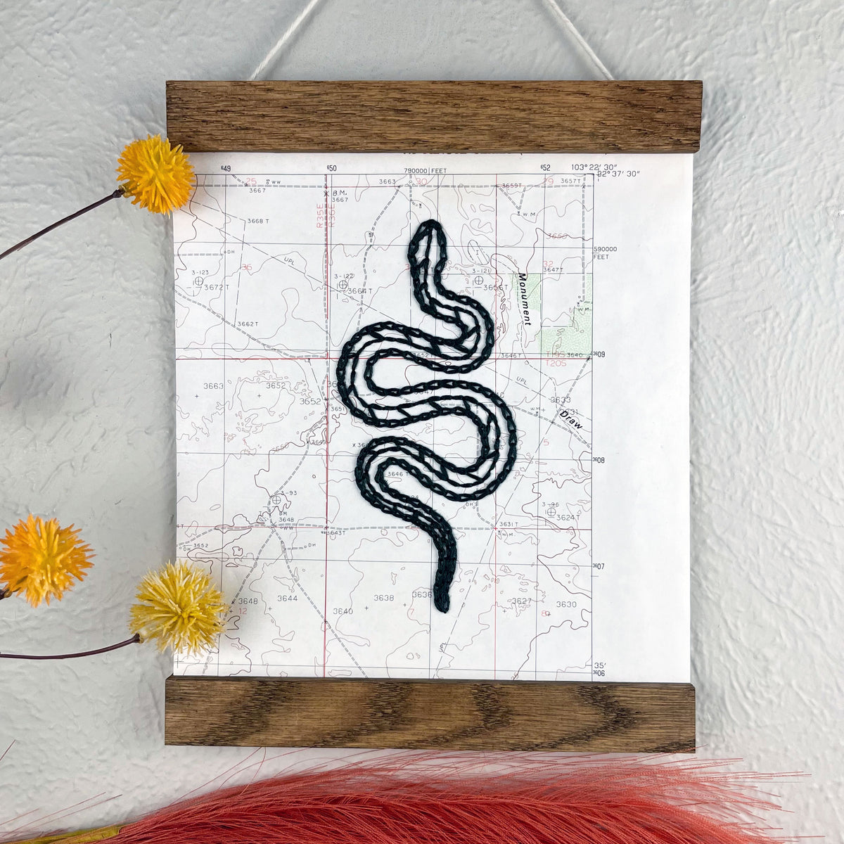 a vintage USGS map of New Mexico hand embroidered with a dark green snake, framed on top and bottom with a rustic wood magnetic frame hanging on a grey wall with  botanical decorations around it