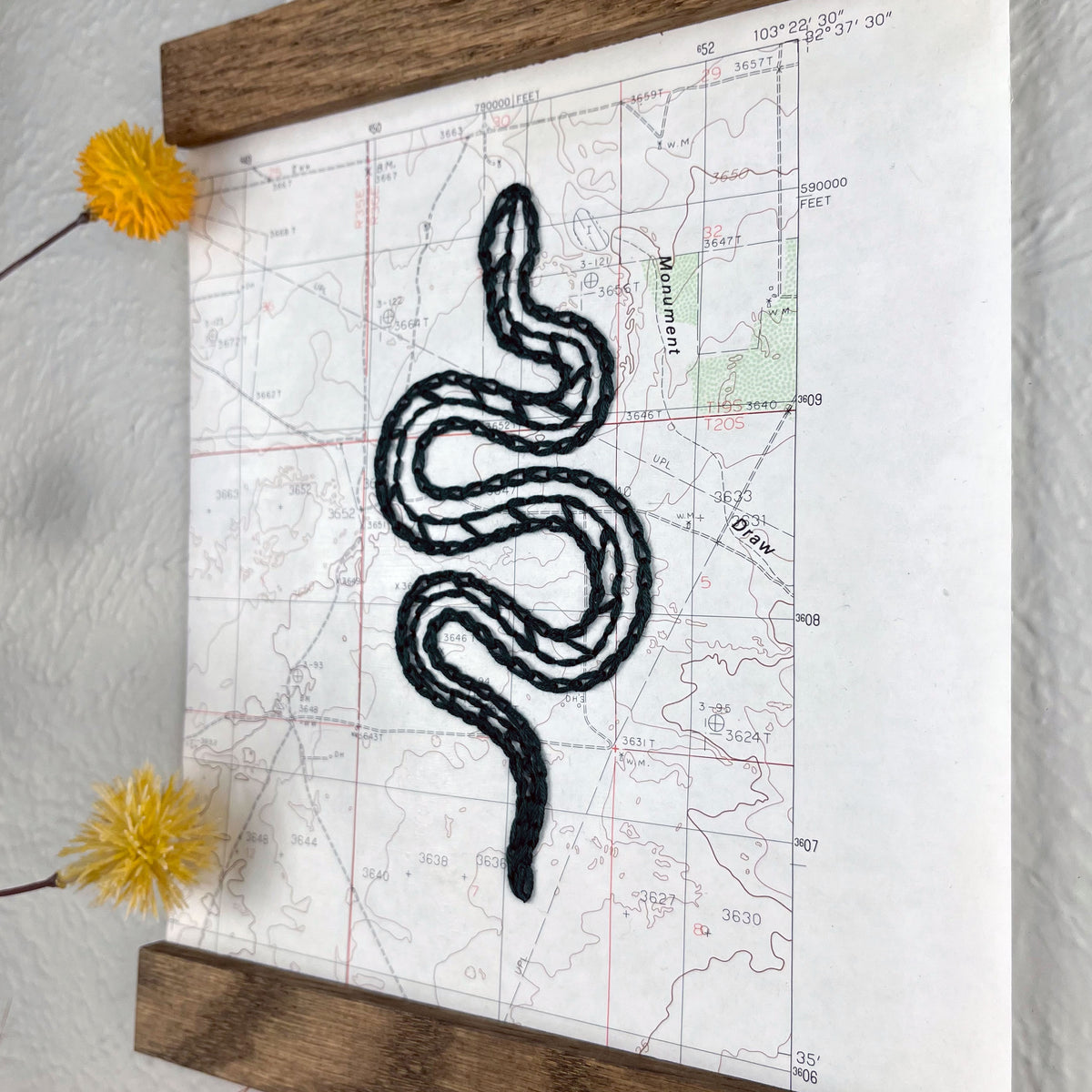 a vintage USGS map of New Mexico hand embroidered with a dark green snake, framed on top and bottom with a rustic wood magnetic frame hanging on a grey wall with  botanical decorations around it