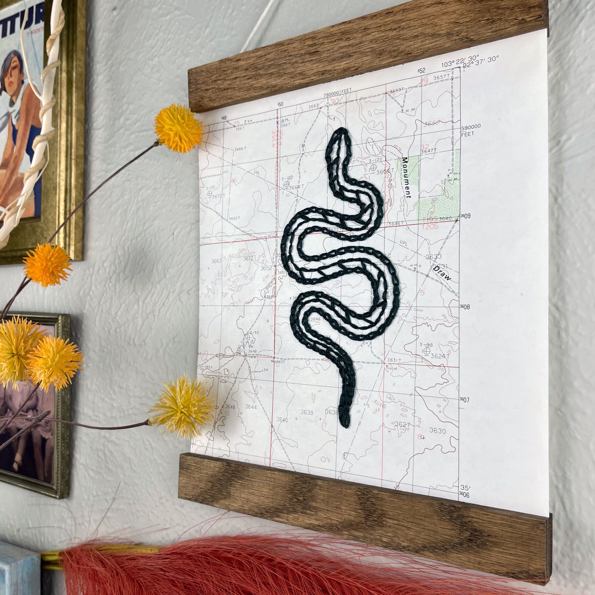 an angled view of a vintage USGS map of New Mexico hand embroidered with a dark green snake, framed on top and bottom with a rustic wood magnetic frame hanging on a grey wall with  framed art and botanical decorations around it