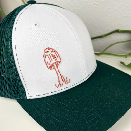 green mesh hat with mauve mushroom hand embroidered to the front, sitting on a white dresser