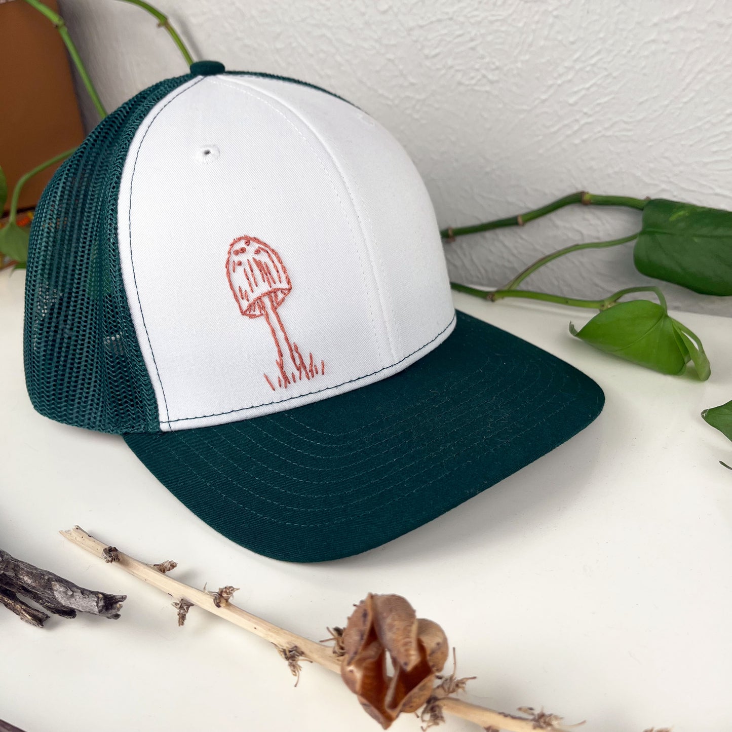 green mesh hat with mauve mushroom hand embroidered to the front, sitting on a white dresser
