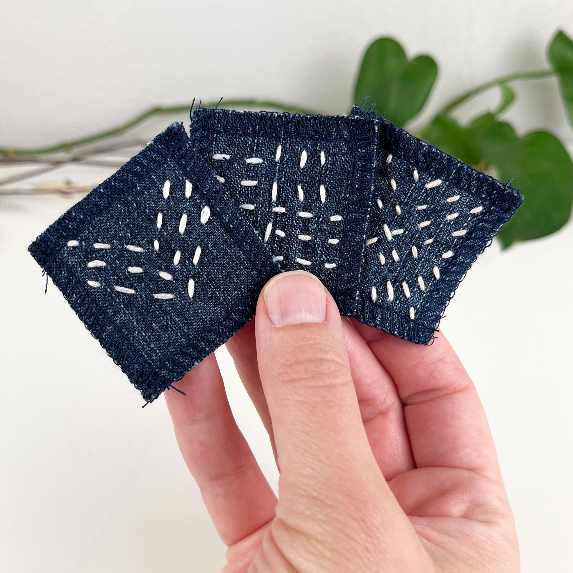 a hand palm up holding three small square denim patches with sashiko style running stitches in ecru of three chevron lines a repeating x pattern  and a basketweave pattern with a white counter and pothos plant in the background 