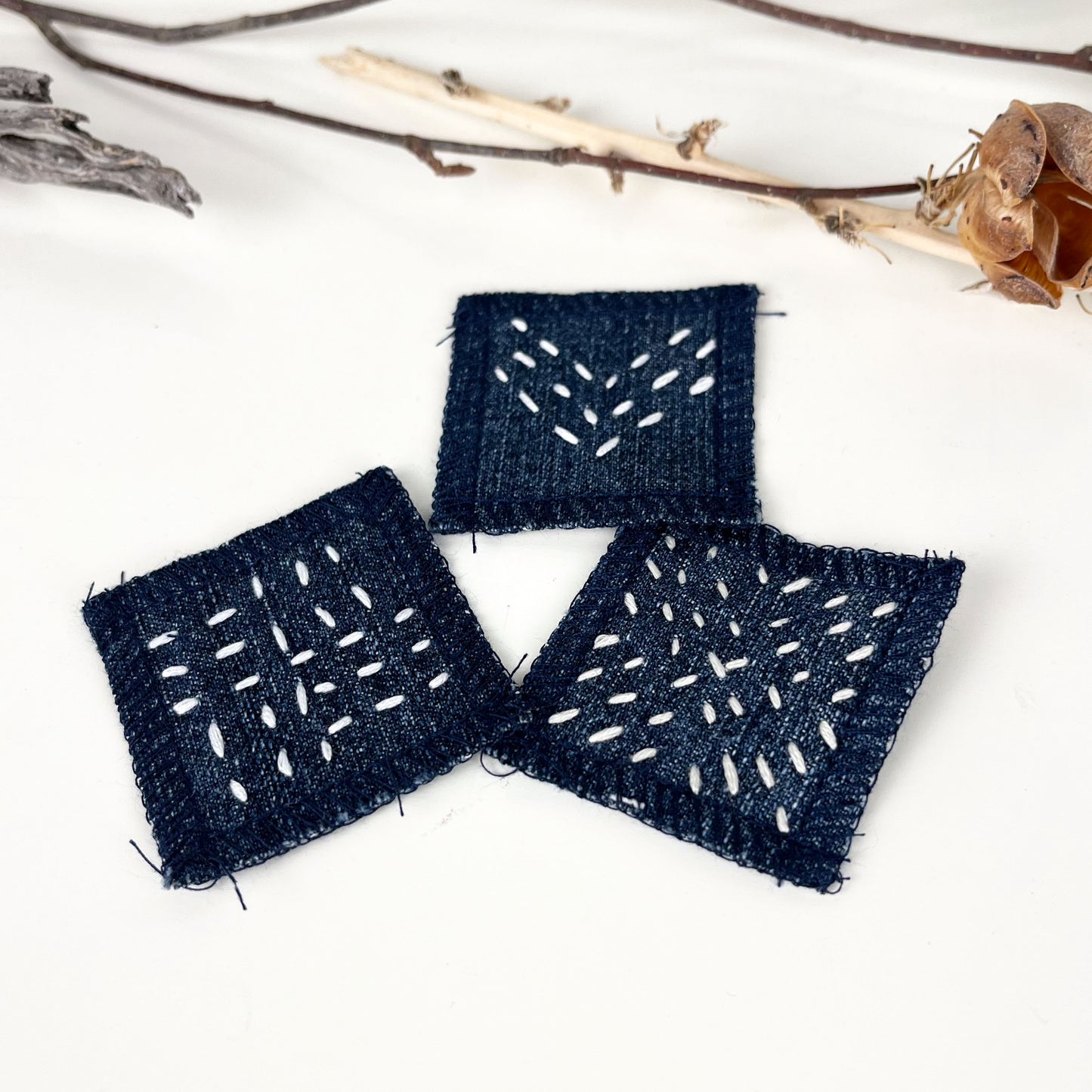  three small square denim patches with sashiko style running stitches in ecru of three chevron lines a repeating x pattern  and a basketweave pattern on a white counter with twigs and a dried seed pod in the background 