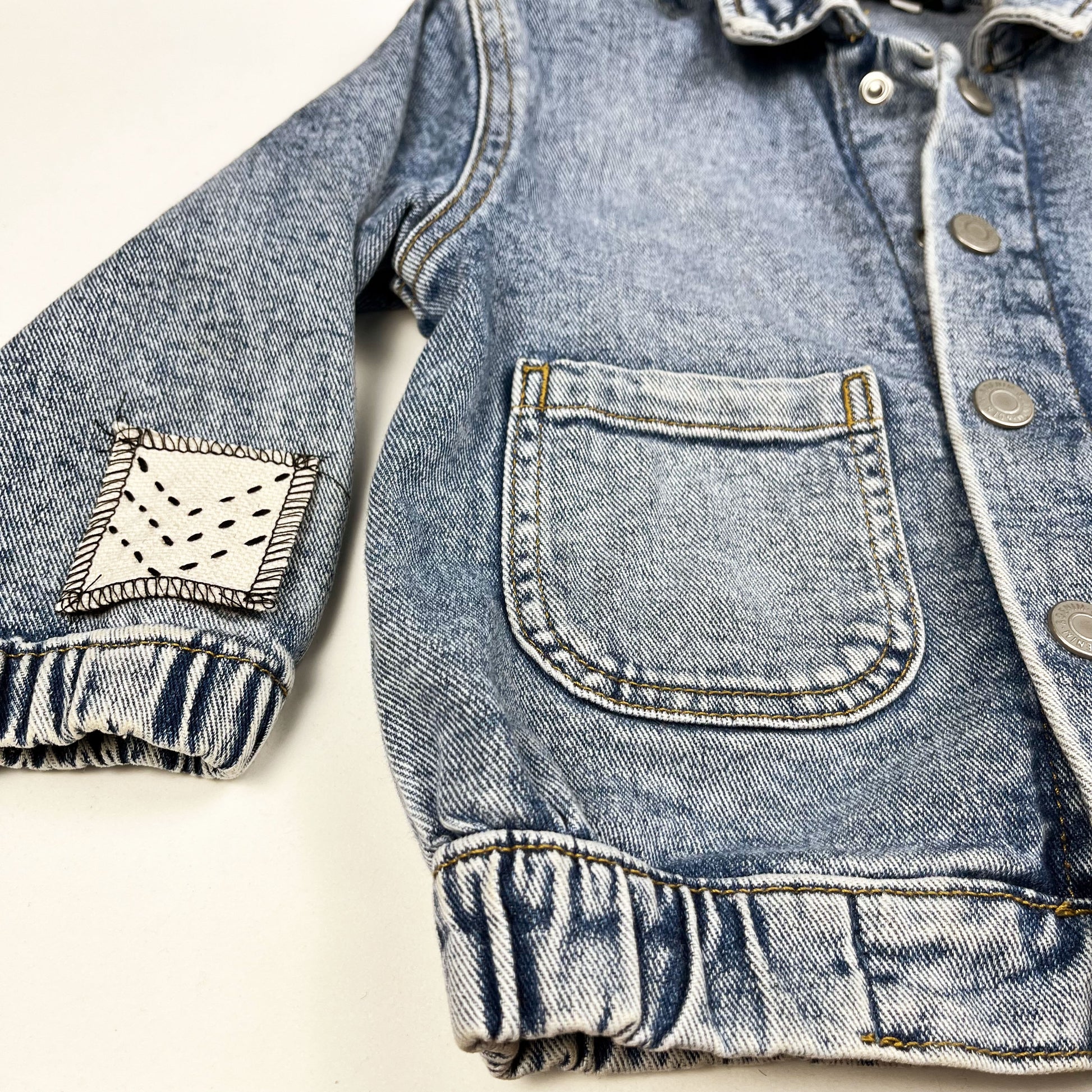angled view of half of a denim jacket, with a small natural colored square patch on the sleeve near the cuff, stitched with three black chevron lines, overlocked in black thread
