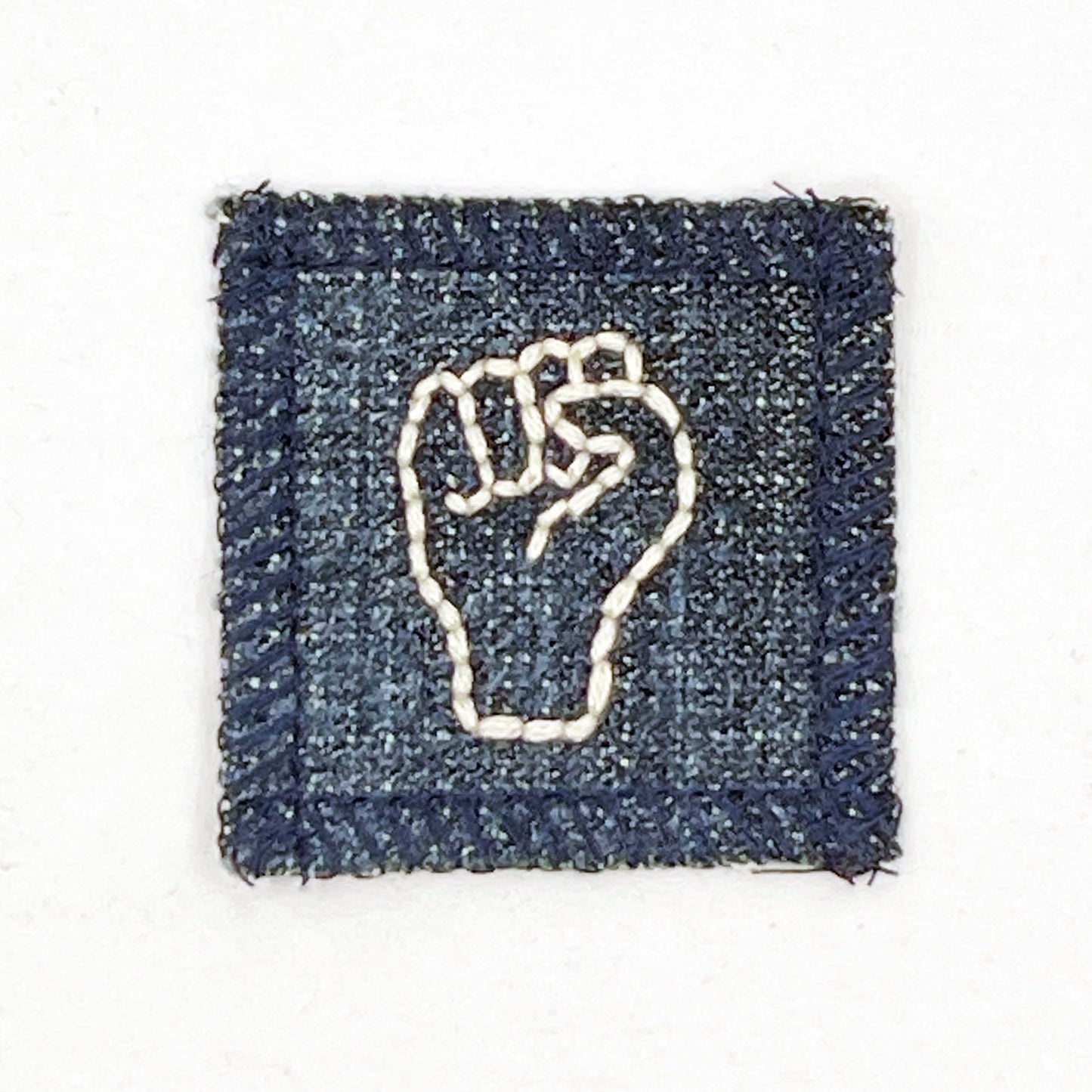 a small denim patch with an empowerment fist symbol hand embroidered in off white thread 