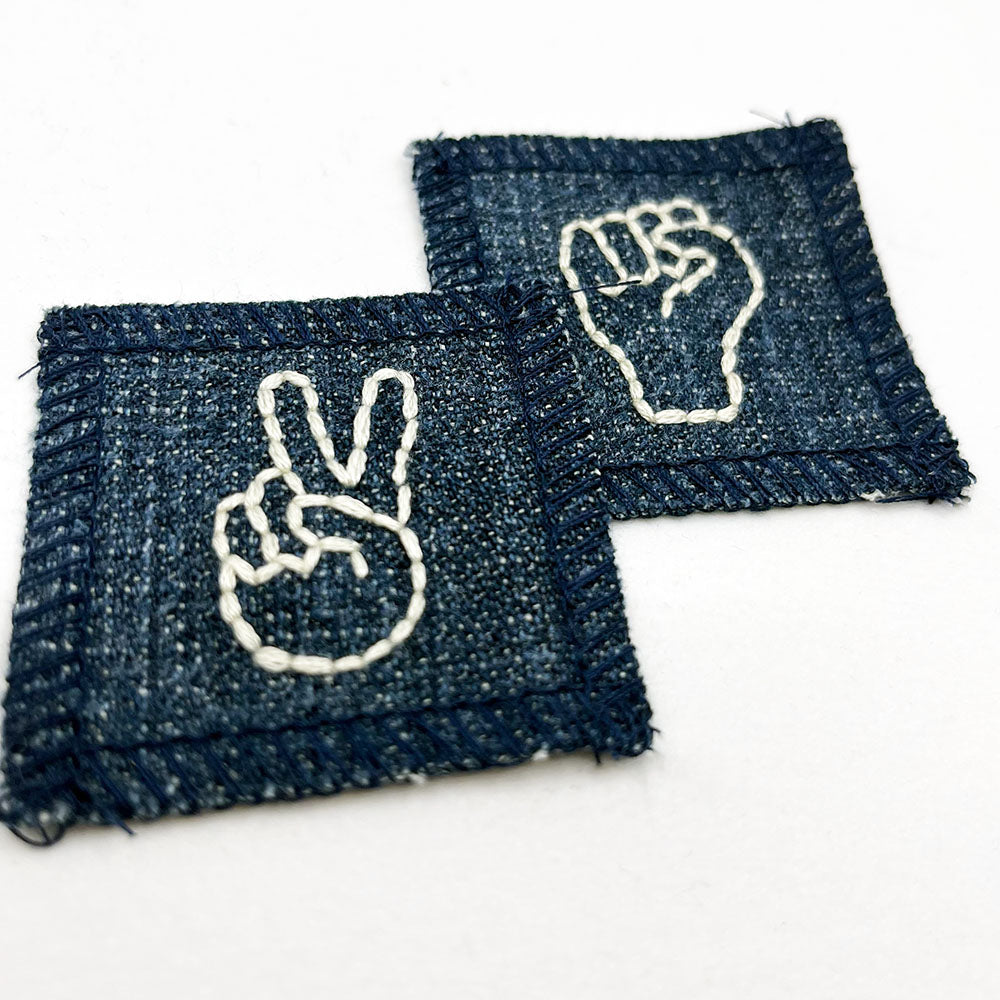 two small denim patches one has a hand embroidered drawing of a hand giving the peace symbol and the other a hand making an empowerment fist