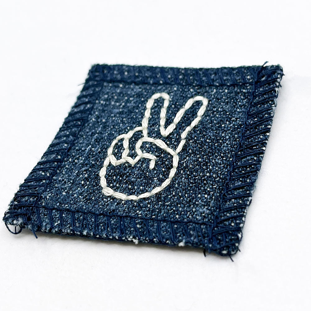 a small denim patch with a hand giving the peace sign hand embroidered in off white thread 