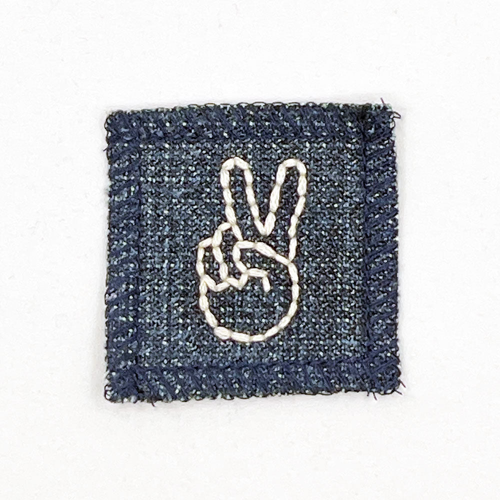 a small denim patch with a hand giving the peace sign hand embroidered in off white thread 