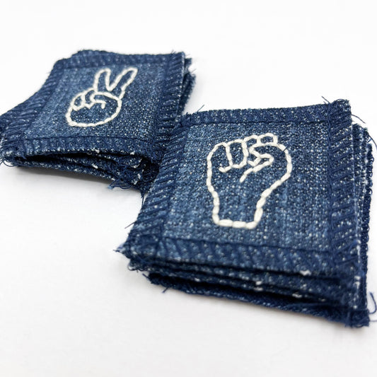 two piles of small denim patches the top of one pile has a hand embroidered drawing of a hand giving the peace symbol and the other a hand making an empowerment fist