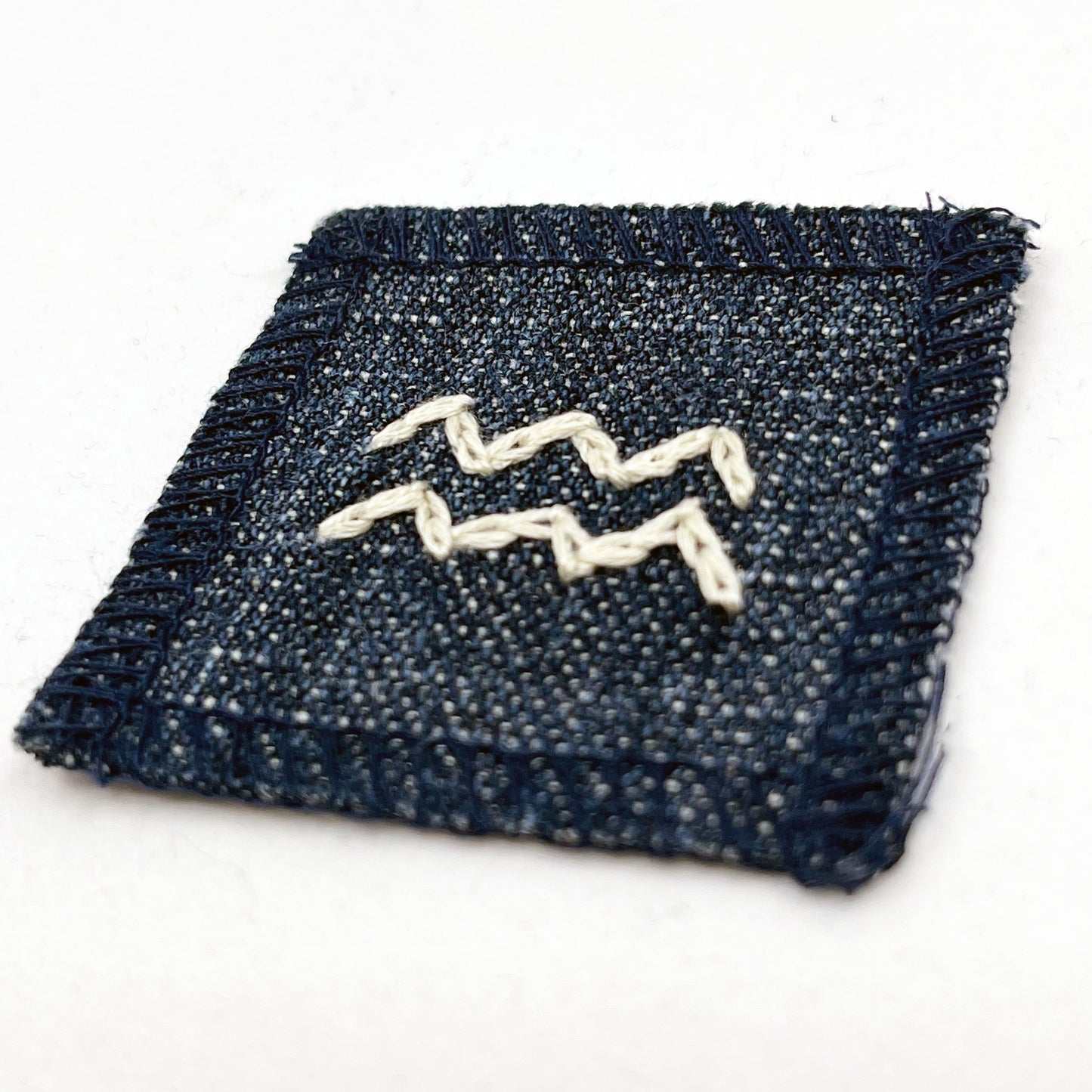 a small square dark denim patch with overlocked edges with the zodiac symbol for aquarius chainstitched on it in off white embroidery floss