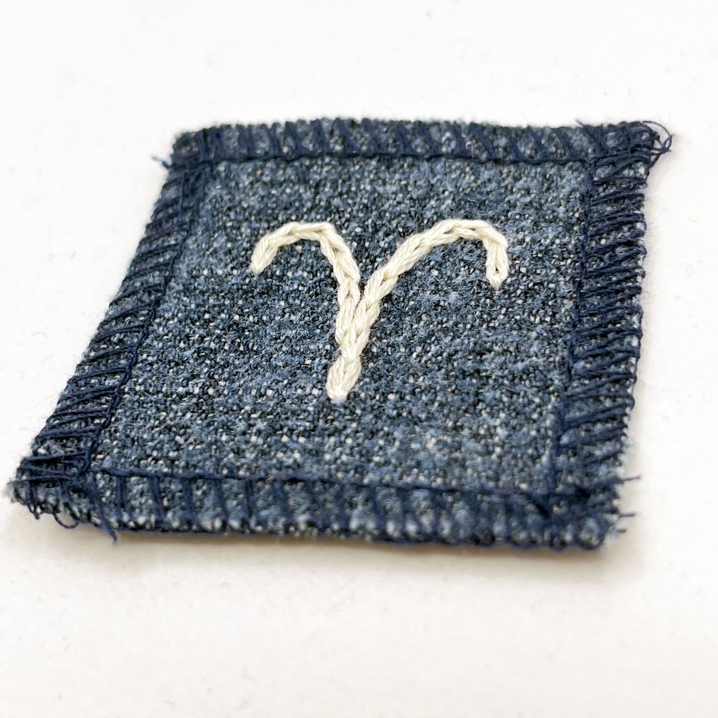 a small square dark denim patch with overlocked edges with the zodiac symbol for aries chainstitched on it in off white embroidery floss