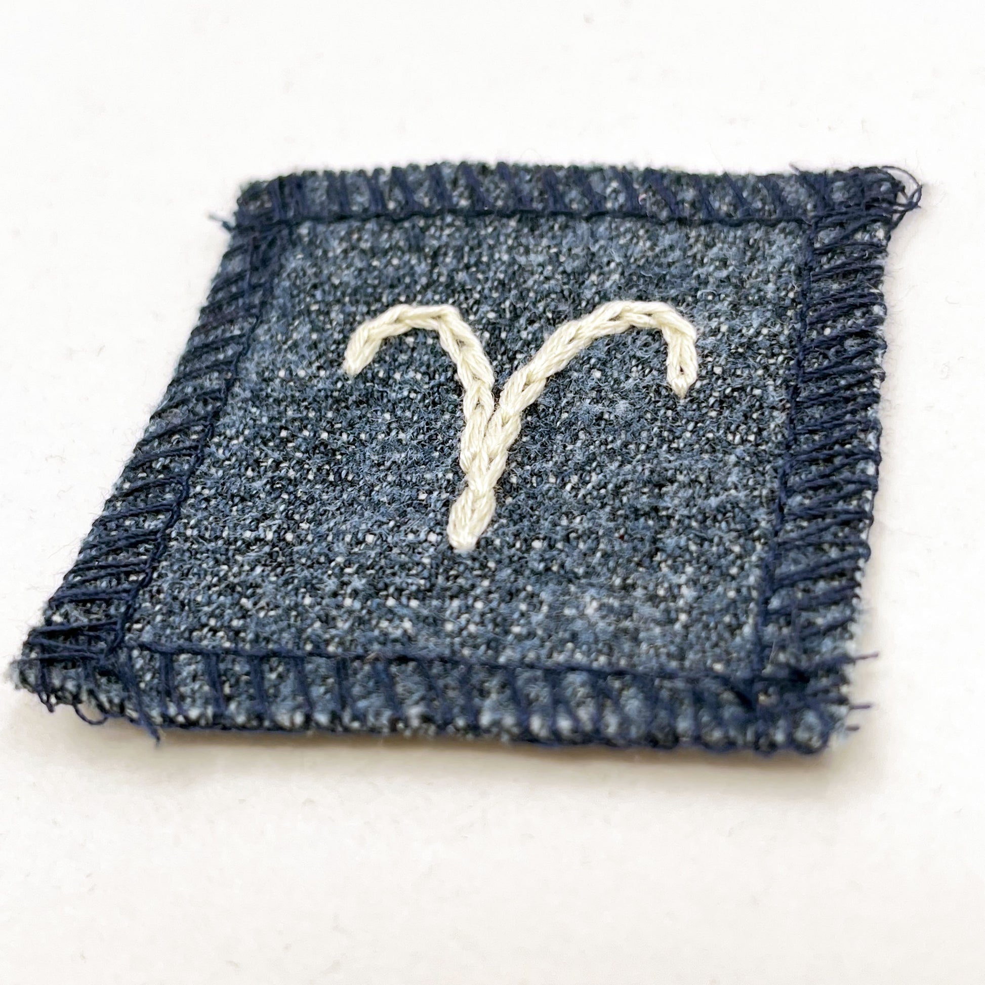 a small square dark denim patch with overlocked edges with the zodiac symbol for aries chainstitched on it in off white embroidery floss
