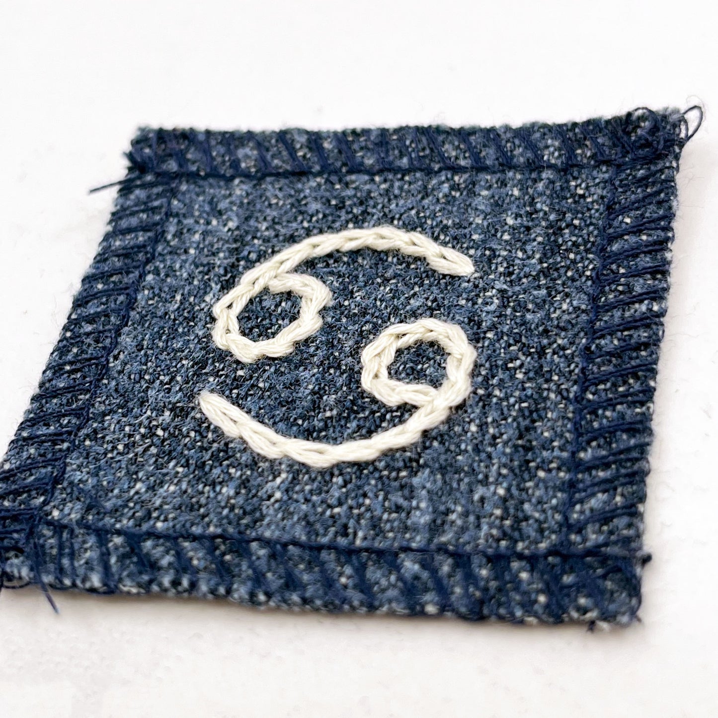 a small square dark denim patch with overlocked edges with the zodiac symbol for cancer chainstitched on it in off white embroidery floss