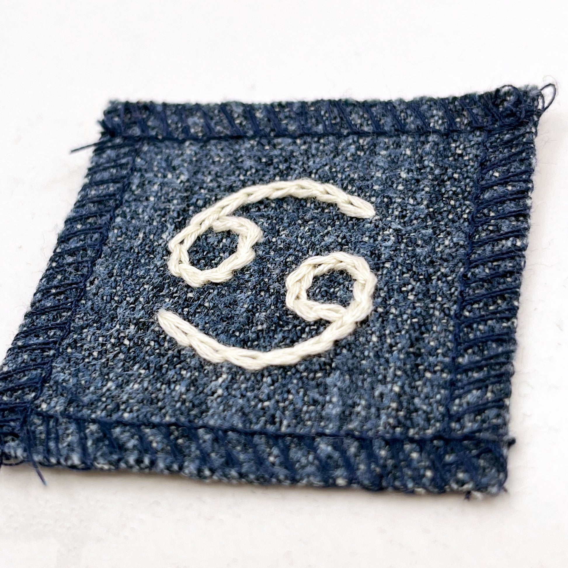 a small square dark denim patch with overlocked edges with the zodiac symbol for cancer chainstitched on it in off white embroidery floss