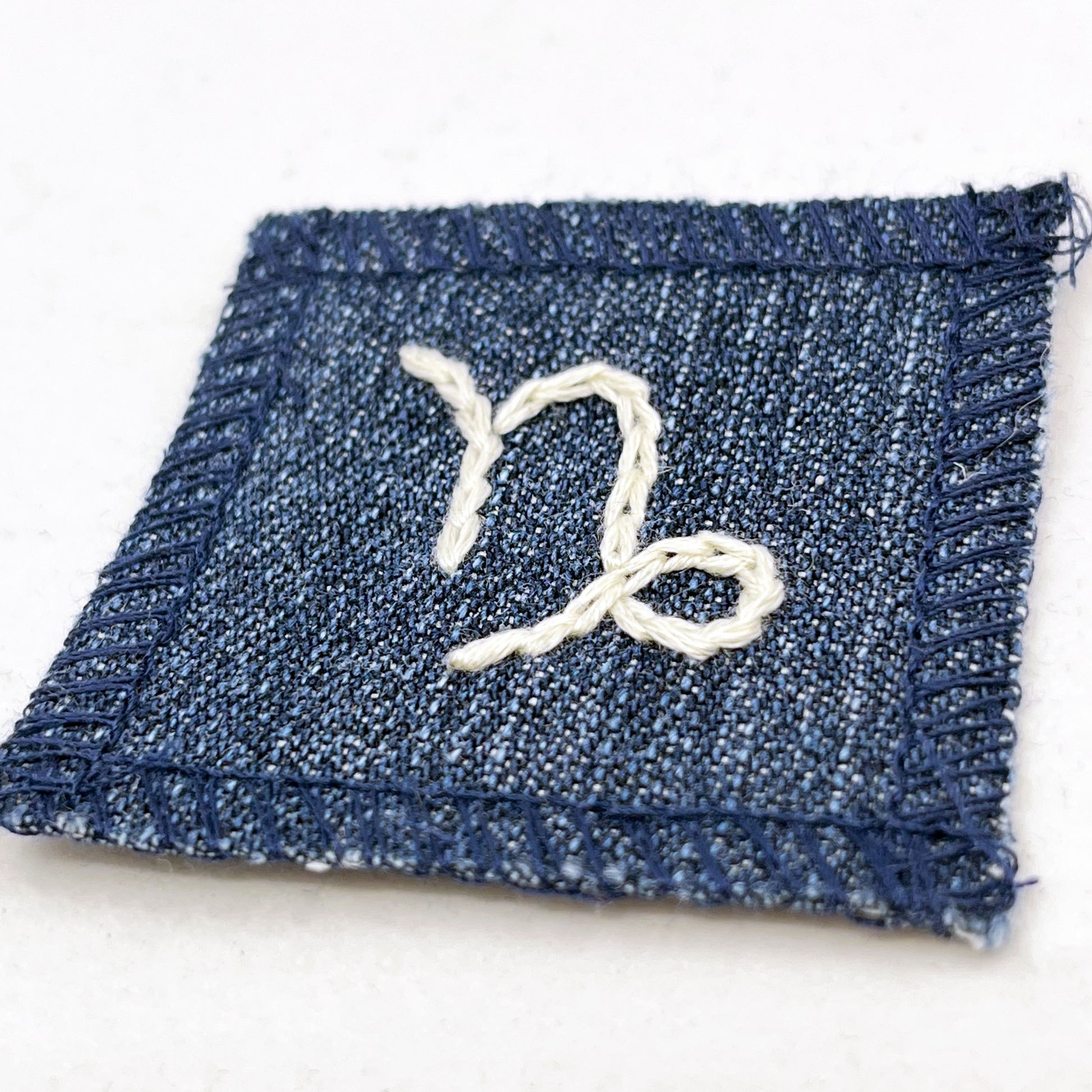 a small square dark denim patch with overlocked edges with the zodiac symbol for capricorn chainstitched on it in off white embroidery floss
