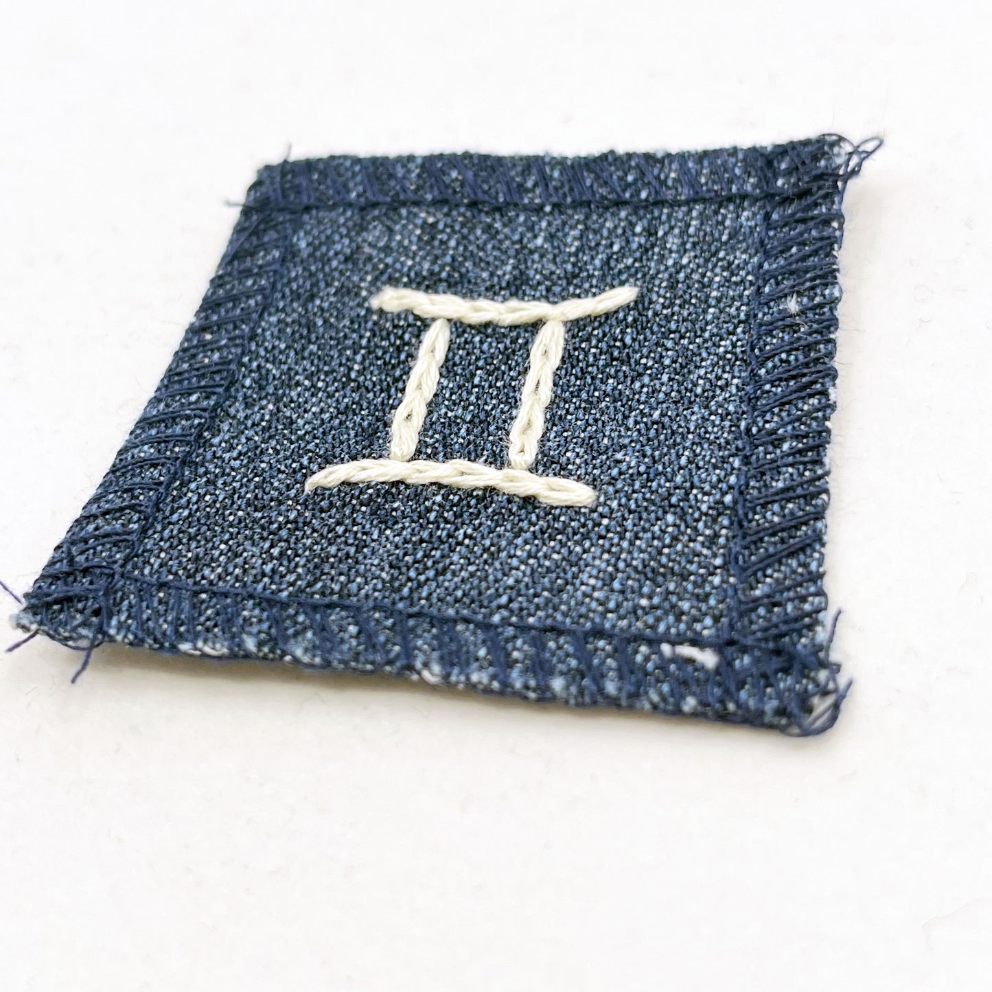 a small square dark denim patch with overlocked edges with the zodiac symbol for gemini chainstitched on it in off white embroidery floss