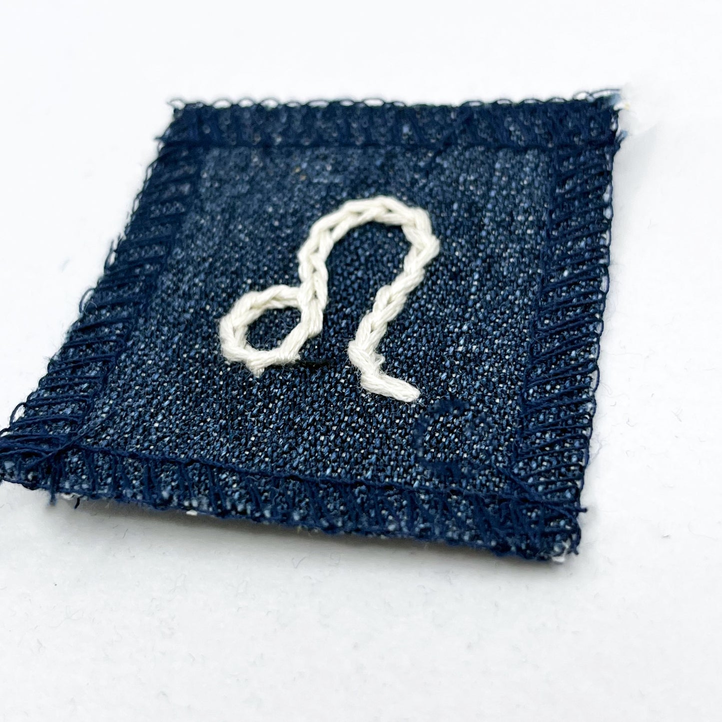 a small square dark denim patch with overlocked edges with the zodiac symbol for leo chainstitched on it in off white embroidery floss
