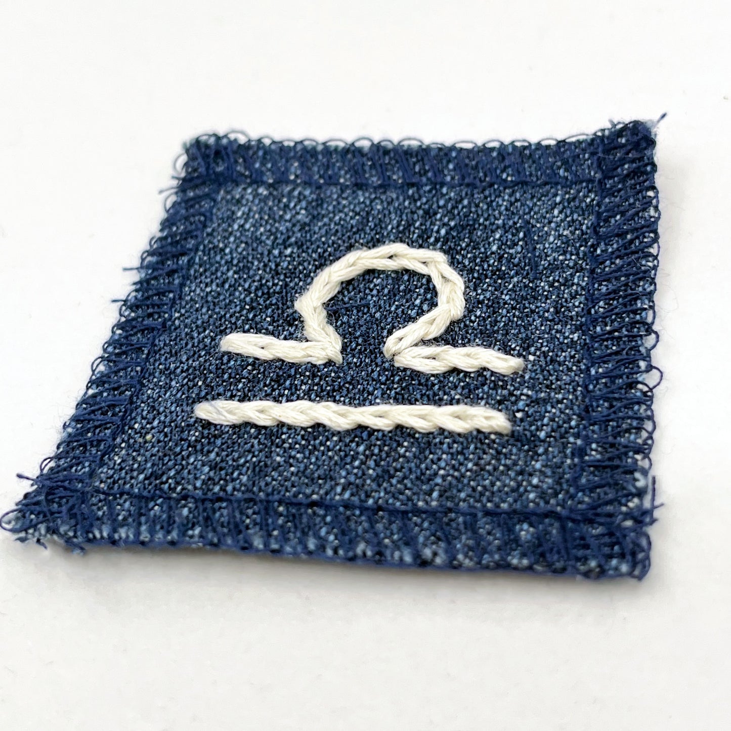 a small square dark denim patch with overlocked edges with the zodiac symbol for libra chainstitched on it in off white embroidery floss