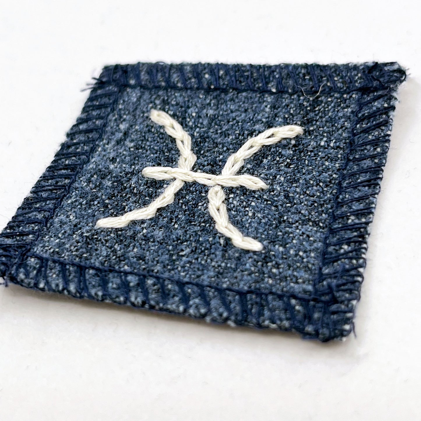 a small square dark denim patch with overlocked edges with the zodiac symbol for pisces chainstitched on it in off white embroidery floss