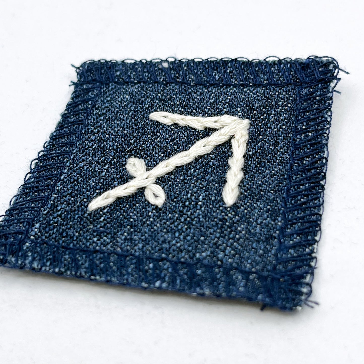 a small square dark denim patch with overlocked edges with the zodiac symbol for sagittarius chainstitched on it in off white embroidery floss