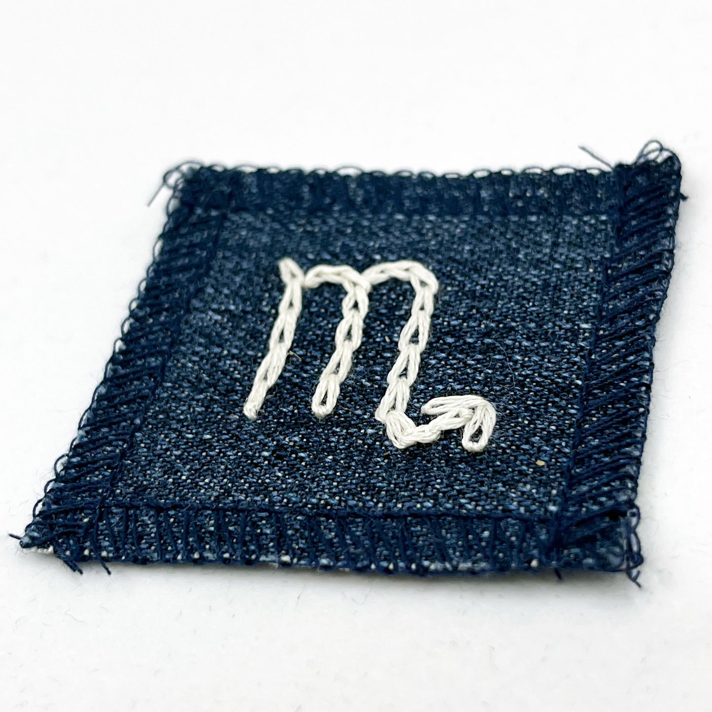 a small square dark denim patch with overlocked edges with the zodiac symbol for scorpio chainstitched on it in off white embroidery floss