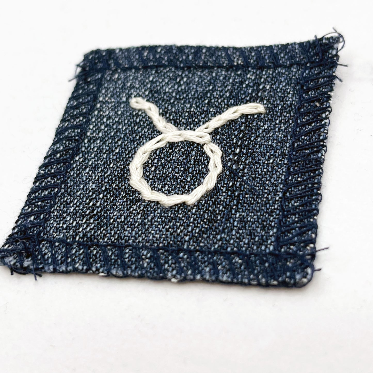a small square dark denim patch with overlocked edges with the zodiac symbol for taurus chainstitched on it in off white embroidery floss