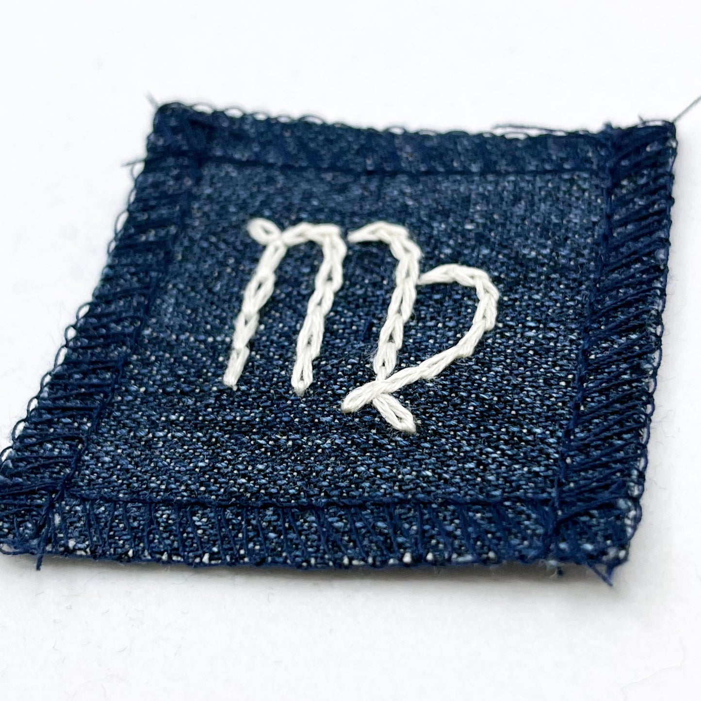 a small square dark denim patch with overlocked edges with the zodiac symbol for virgo chainstitched on it in off white embroidery floss
