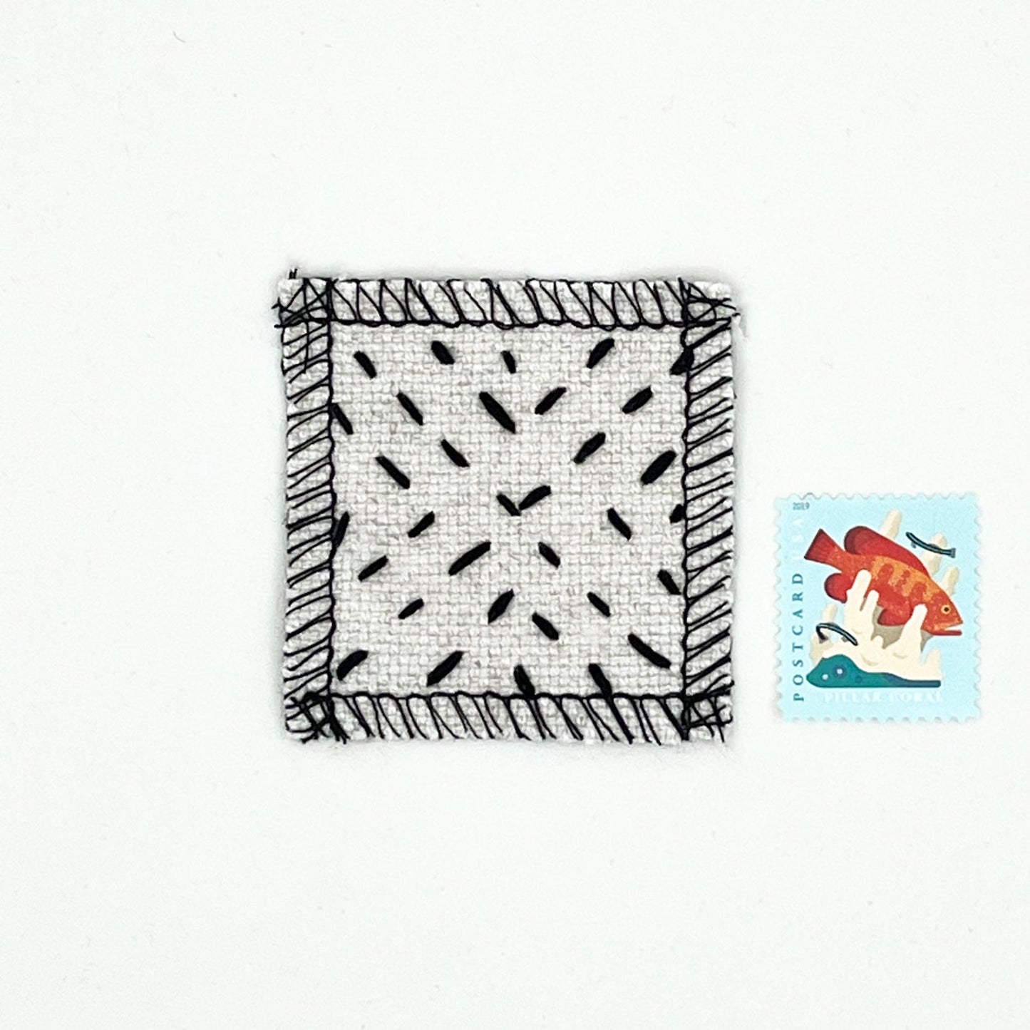 square natural colored patch with stitching in black of a radiating X shape, next to a stamp for size comparison