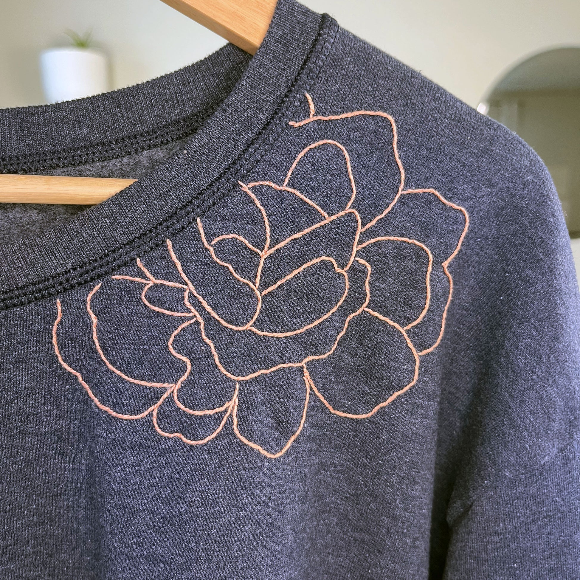 close up view of a navy colored sweatshirt with a peach hand embroidered outline of a peony on the front shoulder and into the front neckline, on a wood hanger