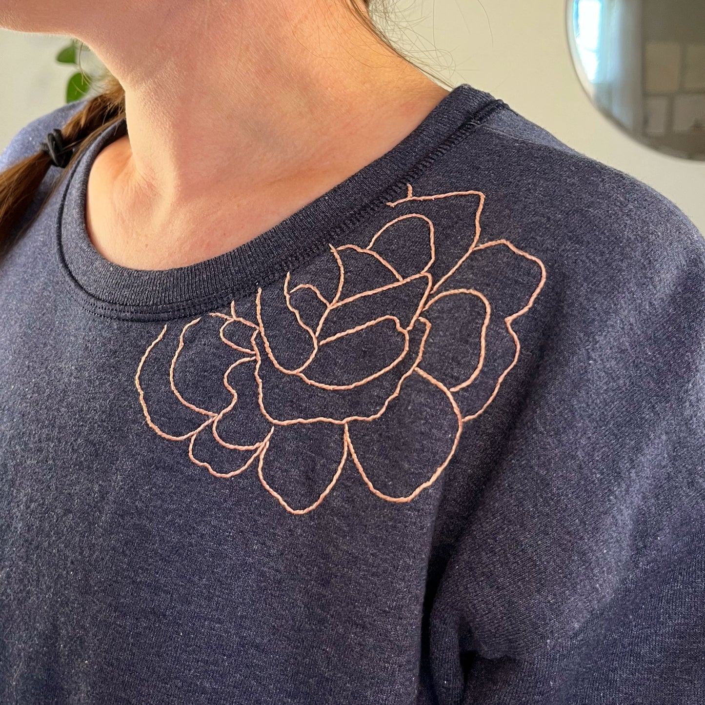 close up view of a woman's shoulder, she's wearing a navy colored sweatshirt with a peach hand embroidered outline of a peony on the front shoulder and into the front neckline
