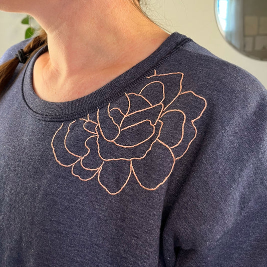 close up view of a woman's shoulder, she's wearing a navy colored sweatshirt with a peach hand embroidered outline of a peony on the front shoulder and into the front neckline