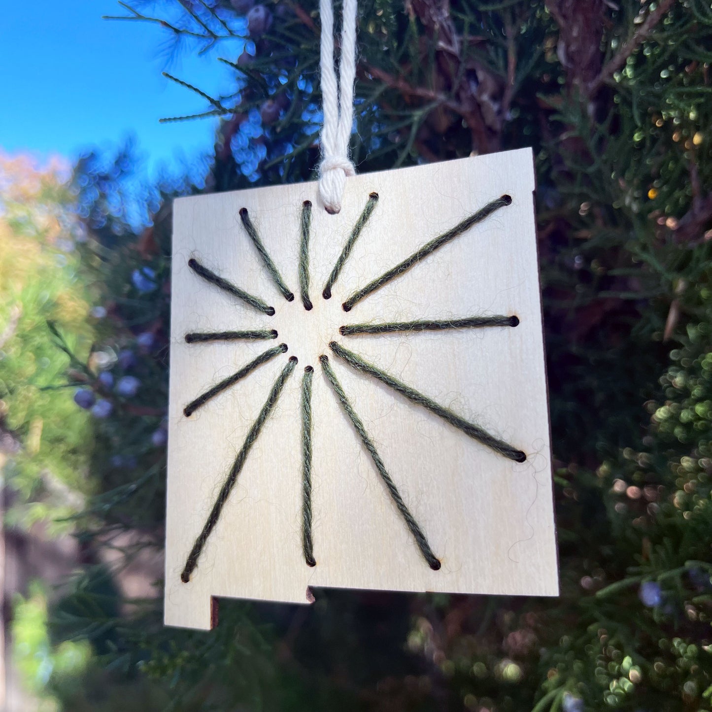 a laser cut wood ornament in the shape of New Mexico, hand embroidered in green thread with sunbeams coming out of a negative space heart, with an off white cord loop hanging on a pine tree