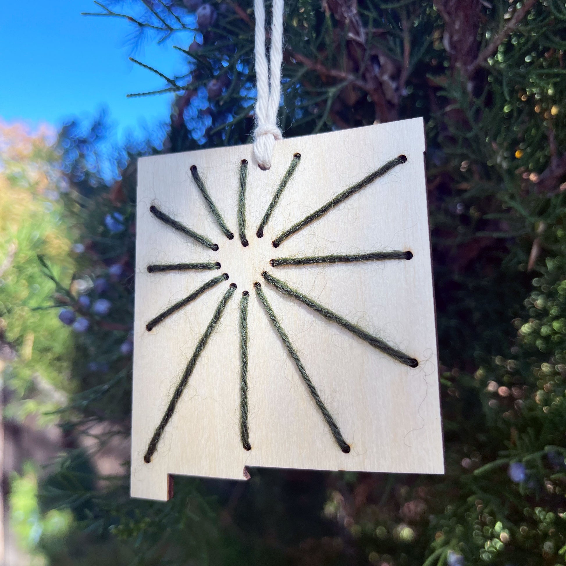 a laser cut wood ornament in the shape of New Mexico, hand embroidered in green thread with sunbeams coming out of a negative space heart, with an off white cord loop hanging on a pine tree