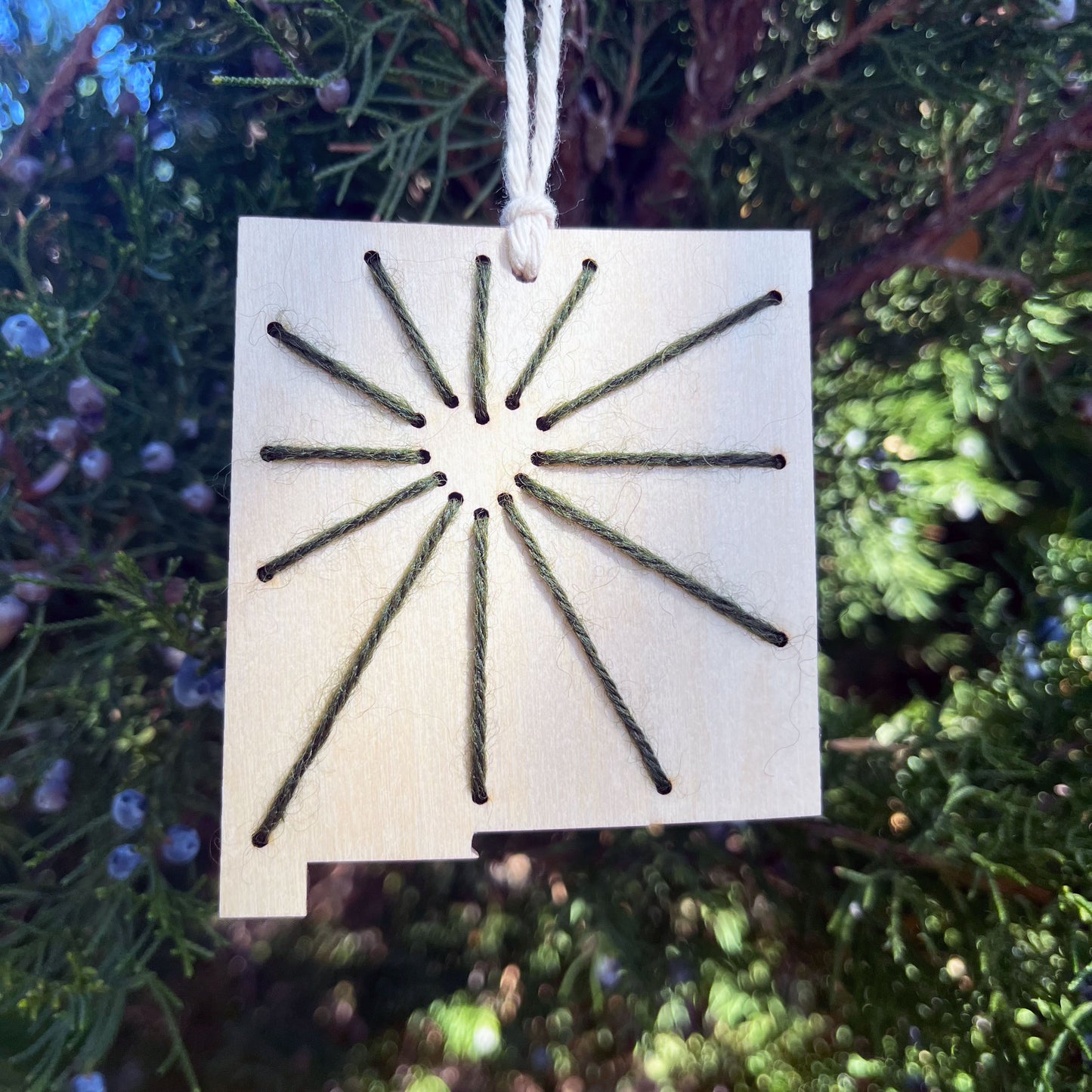 a laser cut wood ornament in the shape of New Mexico, hand embroidered in green thread with sunbeams coming out of a negative space heart, with an off white cord loop hanging on a pine tree