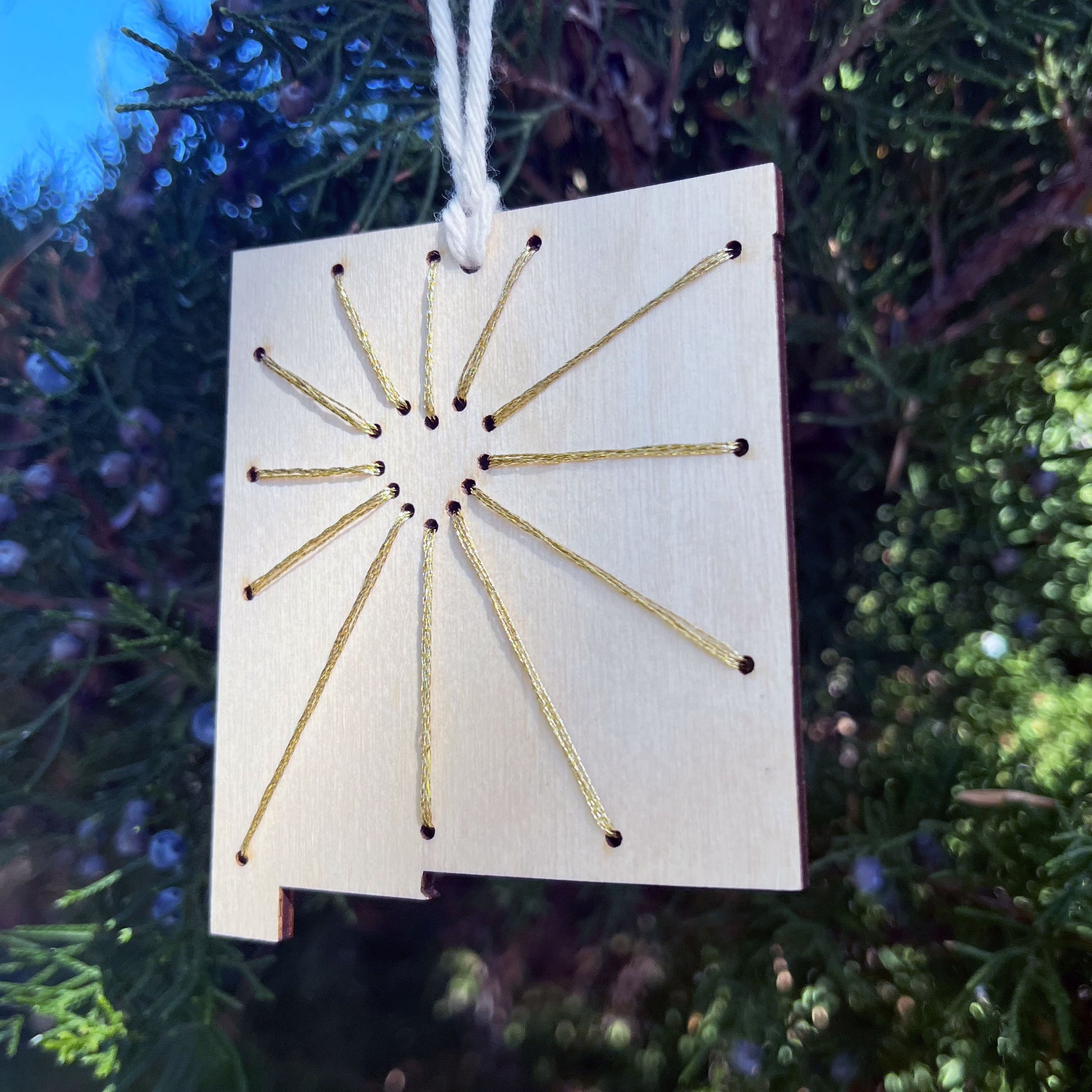 a laser cut wood ornament in the shape of New Mexico, hand embroidered in metallic gold thread with sunbeams coming out of a negative space heart, with an off white cord loop hanging on a pine tree