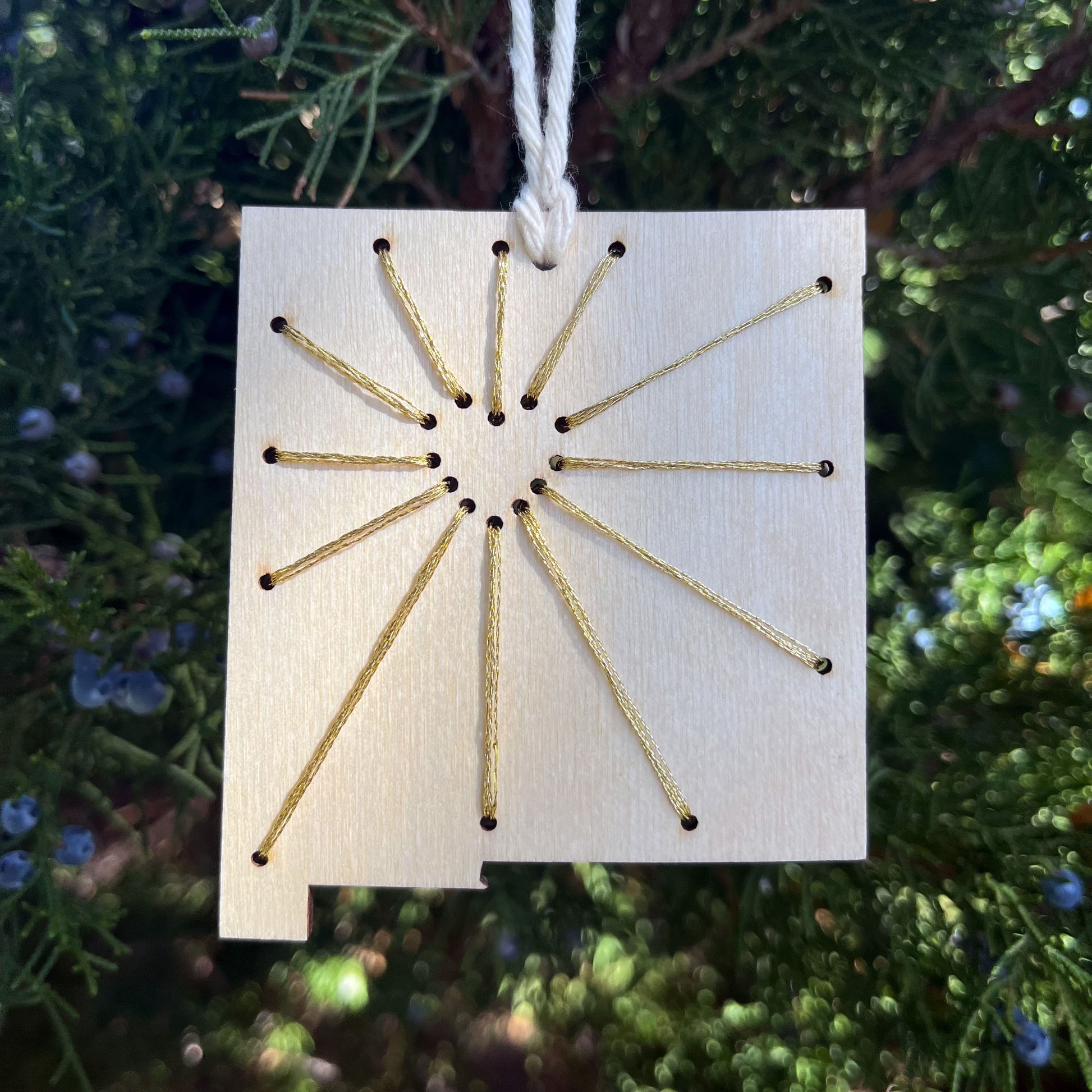 a laser cut wood ornament in the shape of New Mexico, hand embroidered in metallic gold thread with sunbeams coming out of a negative space heart, with an off white cord loop hanging on a pine tree