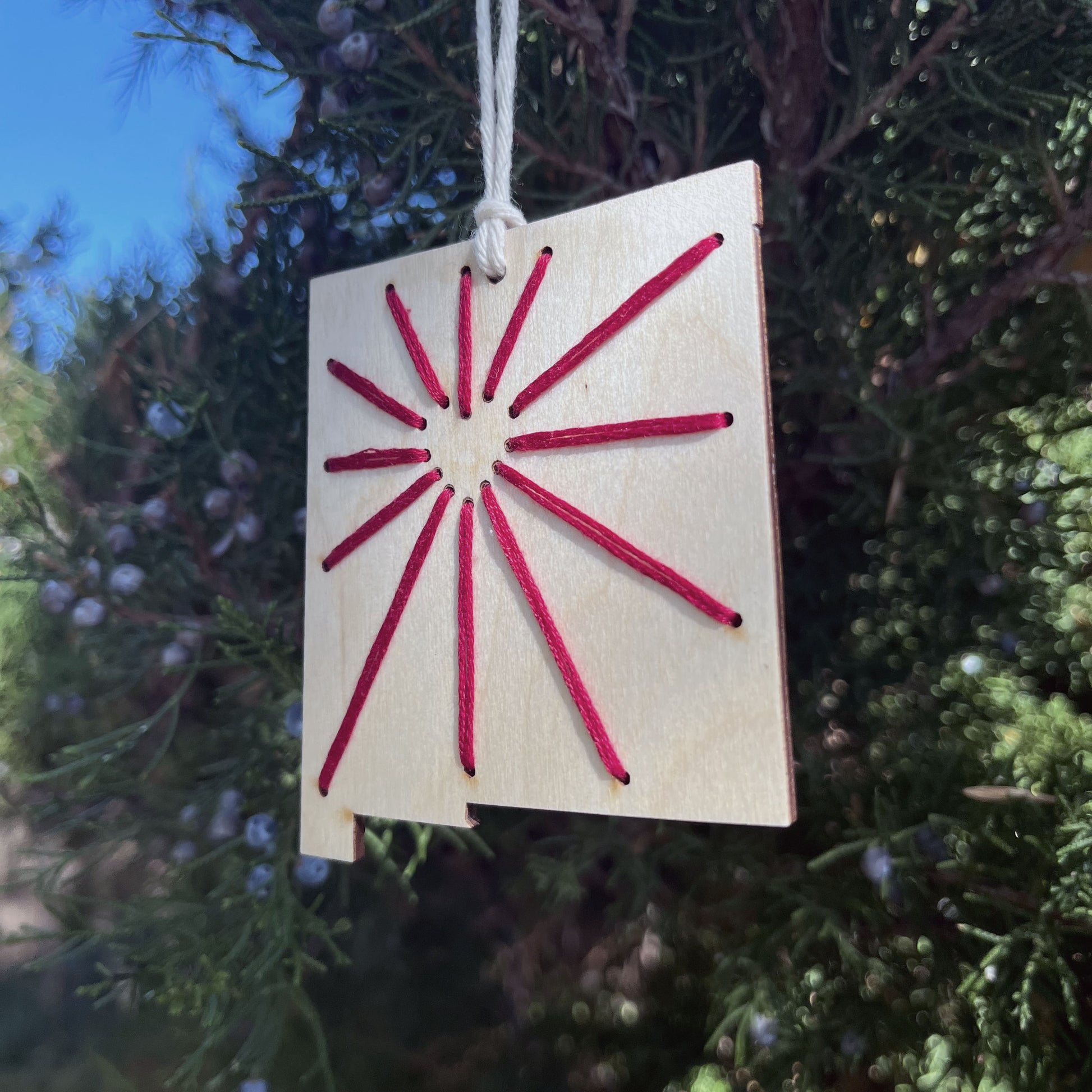 a laser cut wood ornament in the shape of New Mexico, hand embroidered in red thread with sunbeams coming out of a negative space heart, with an off white cord loop hanging on a pine tree
