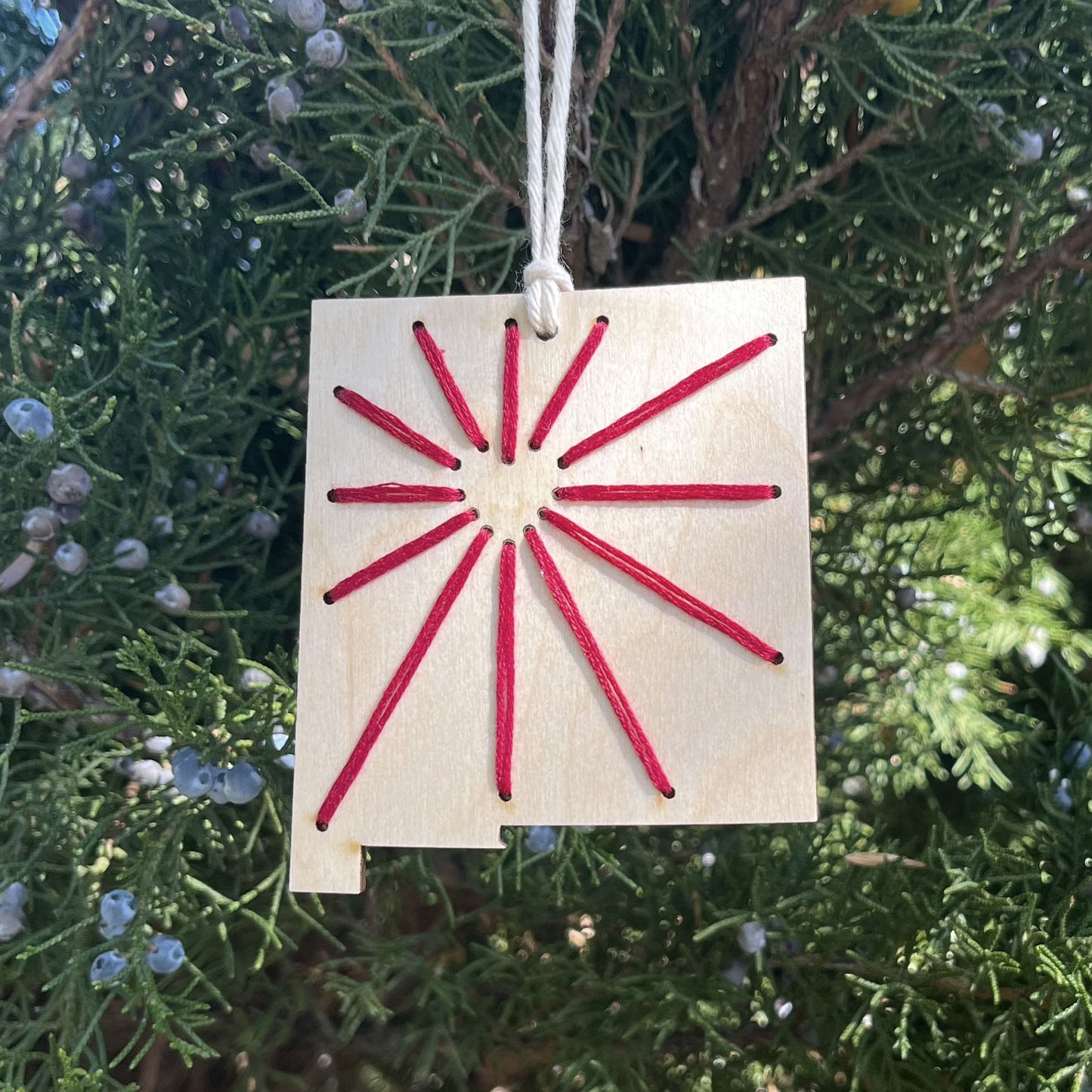 a laser cut wood ornament in the shape of New Mexico, hand embroidered in red thread with sunbeams coming out of a negative space heart, with an off white cord loop hanging on a pine tree