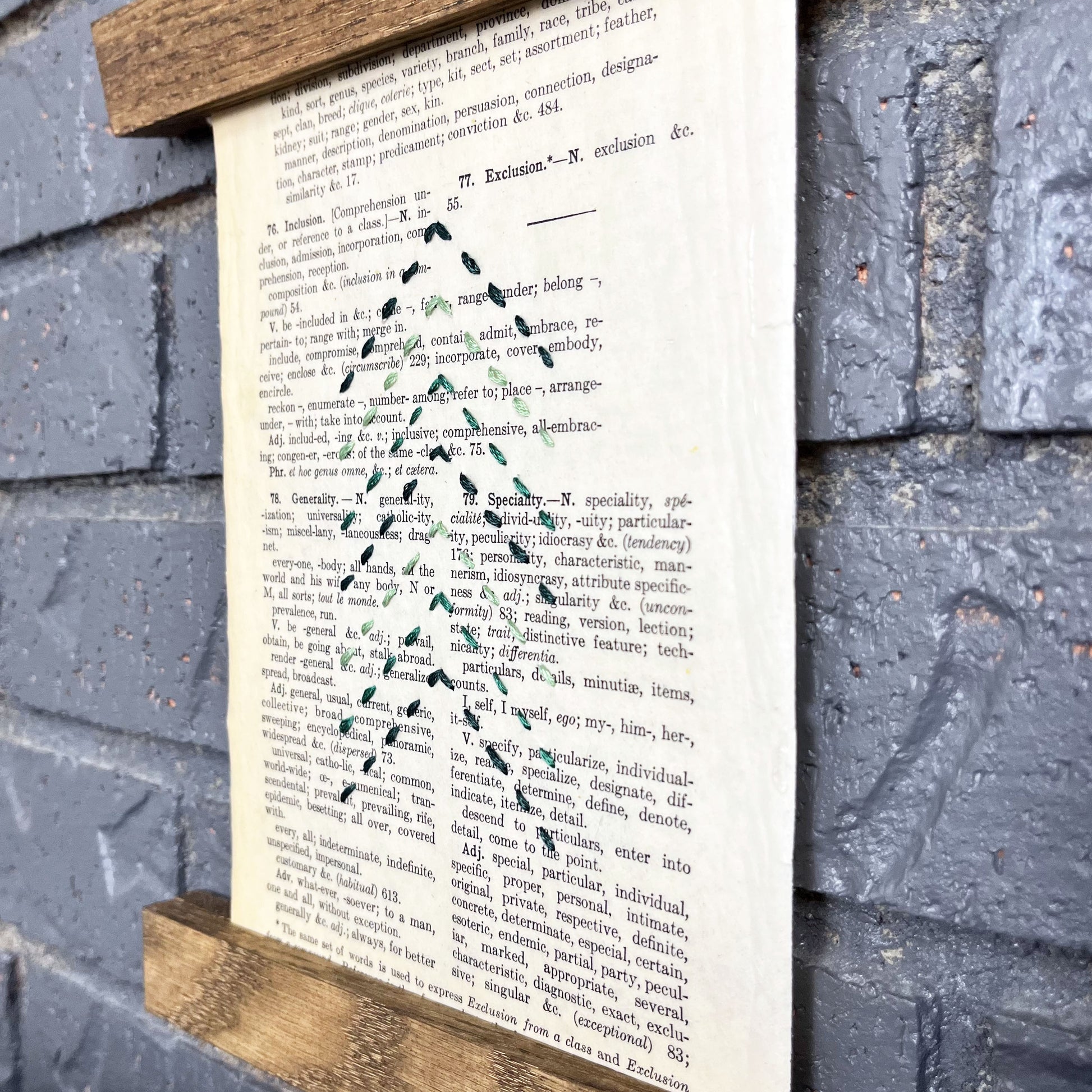 a paper wall hanging in a magnetic wood frame, made from a vintage thesaurus page, embroidered with the shape of a Christmas tree made from running stitches forming a column of chevrons, in shades of pine green thread, hanging on a grey brick wall