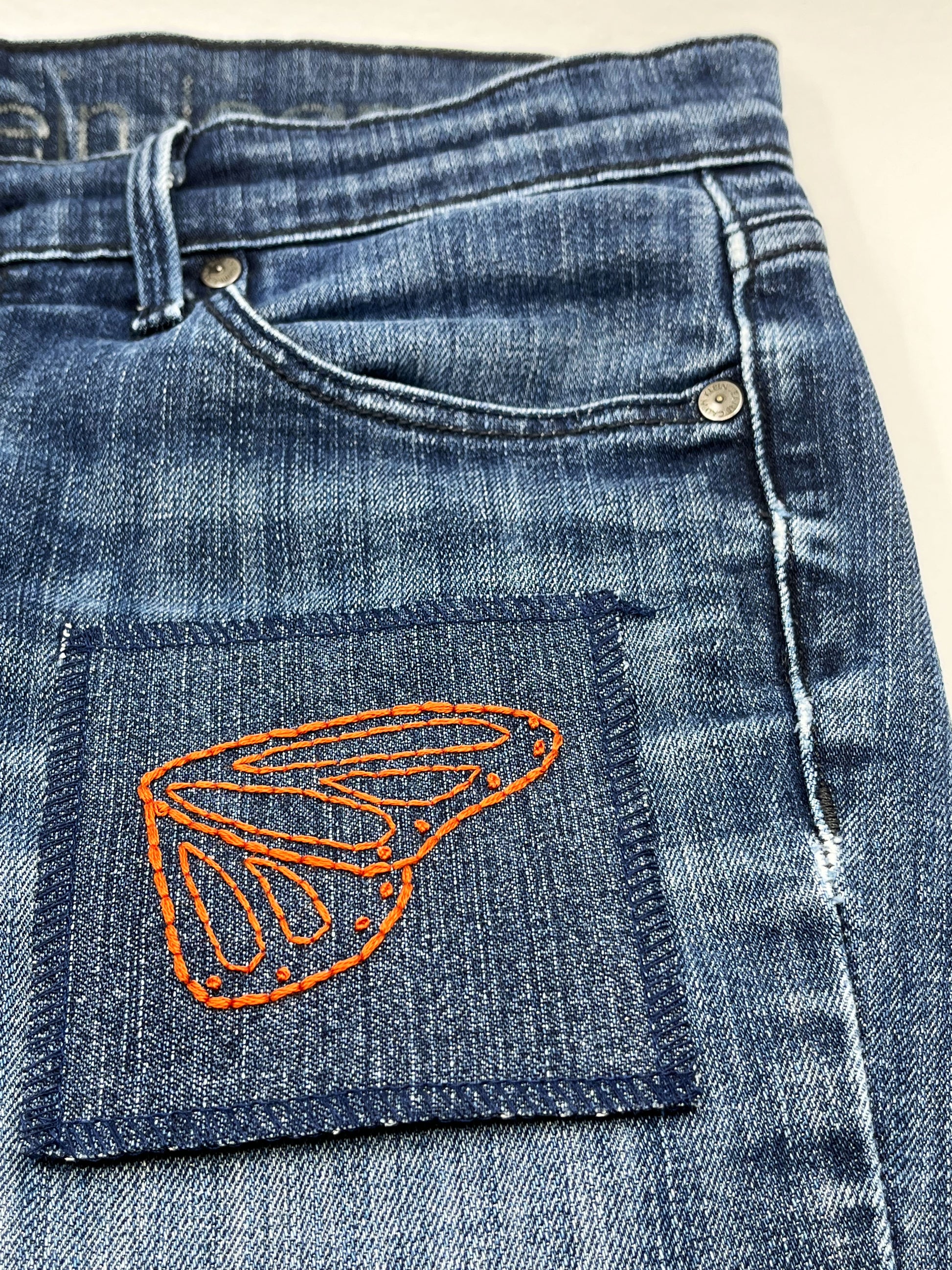 a square denim patch with overlocked edges, with a butterfly wing hand embroidered on it, in rust colored thread with backstitch and french knots, on a pair of jeans