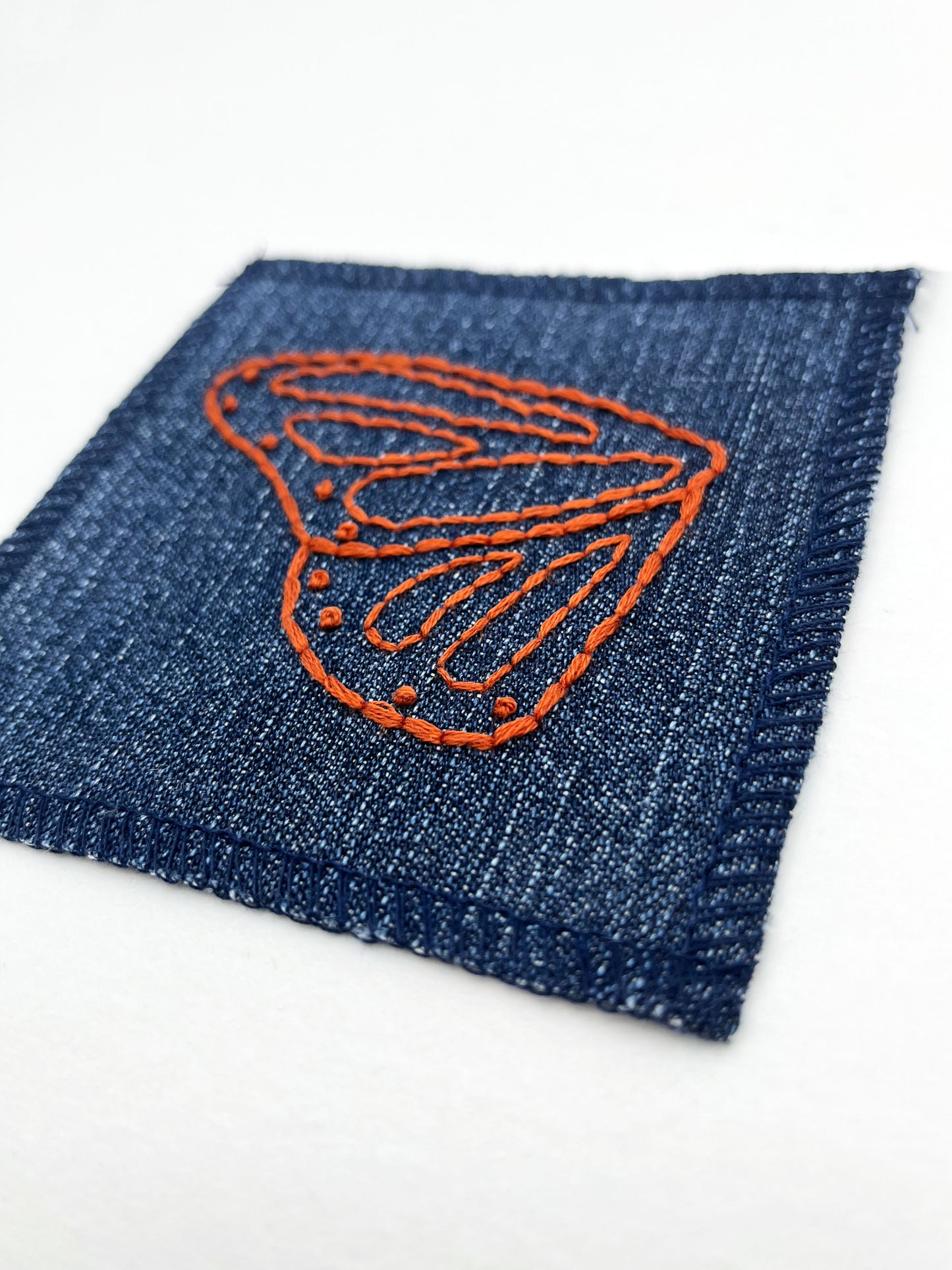 close up angled view of a square denim patch with overlocked edges, with a butterfly wing hand embroidered on it, in rust thread with backstitch and french knots, on a white background