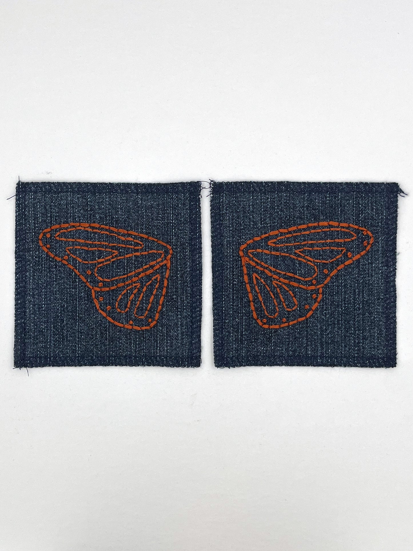 two square denim patches with overlocked edges, with butterfly wings hand embroidered on them, one patch has the left wing and the other has the right wing. in rust thread with backstitch and french knots