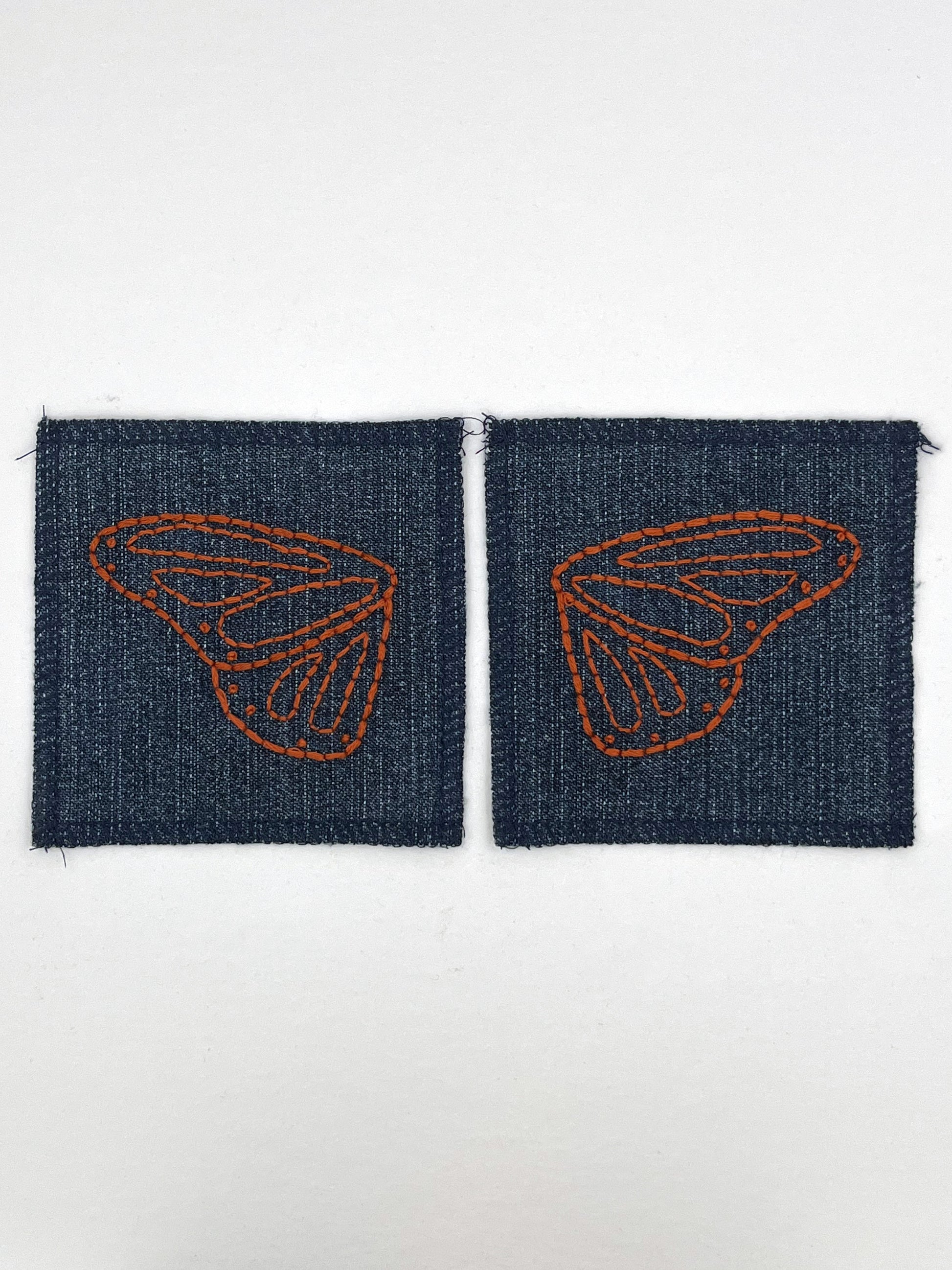 two square denim patches with overlocked edges, with butterfly wings hand embroidered on them, one patch has the left wing and the other has the right wing. in rust thread with backstitch and french knots