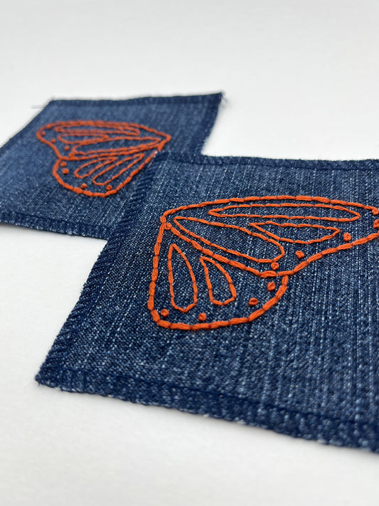 two square denim patches with overlocked edges, with butterfly wings hand embroidered on them, one patch has the left wing and the other has the right wing. in rust thread with backstitch and french knots