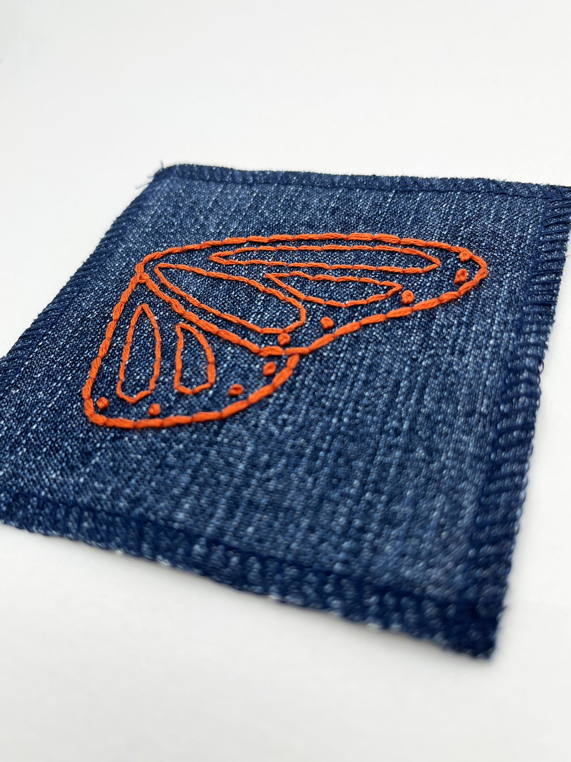 close up angled view of a square denim patch with overlocked edges, with a butterfly wing hand embroidered on it, in rust thread with backstitch and french knots, on a white background