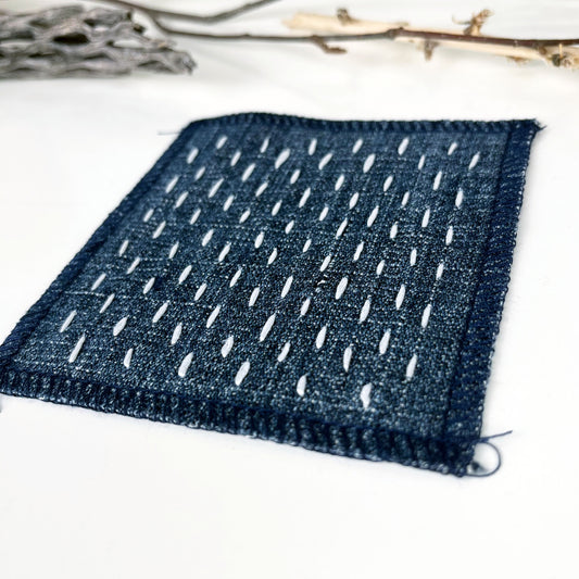 square denim patch with sashiko style running stitches in ivory on a white counter with dried twigs in the background
